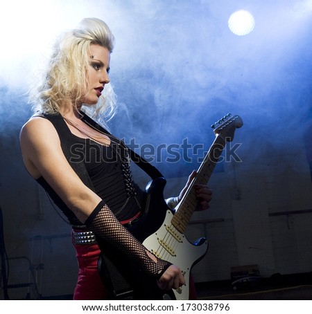 An attractive young woman plays guitar on a stage with lights shining through smoke behind her./Female guitarist