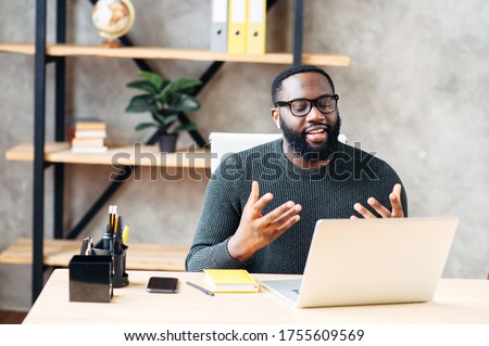 Video call. Attractive African-American guy is using laptop for video communication, a guy in glasses sits at the table in modern office, looking into webcam and talking online