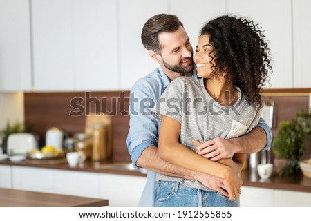 Smiling man hugging from behind charming African American woman, two people standing and joyfully looking at each other. Young international couple happily spending time in cozy modern kitchen at home