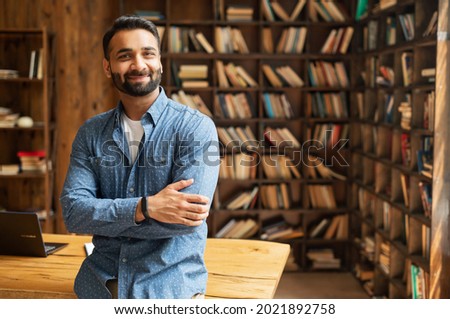 Smiling bearded indian businessman stands near desk and looks at the camera. Young positive male student in library with bookshelves on background. Proud and successful mixed-race small business owner
