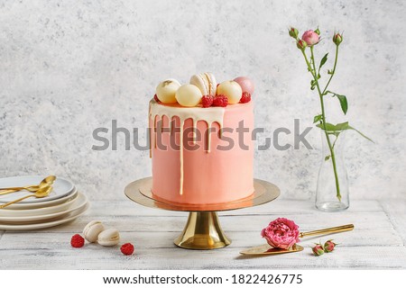 Tall pink cake decorated with macaroons, raspberries and chocolate balls on golden cake stand over white background with flowers and berries. Side view, copy space