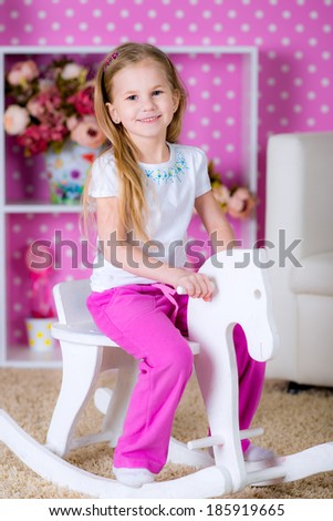 Happy little blonde  girl riding on white wooden horse indoors in a beautiful room with pink background