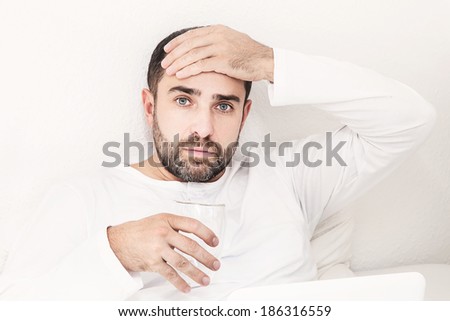Man with glass of water lying on the bed holding his head. Headache.
