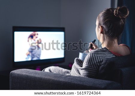 Woman watching romantic movie and eating ice cream. Sad lonely single girl streaming series or film on tv home at night. Person with stream on television. Comfy candid relaxing under blanket on couch.