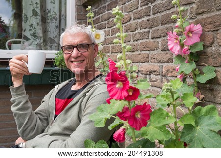 senior man drinks coffee in garden with hollyhocks