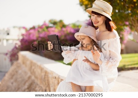 Portrait of happy beautiful caucasian woman and her daughter having videochat while using digital tablet while sitting on the background of tree with flowers and sunny summer landscape, waving hand Foto stock © 