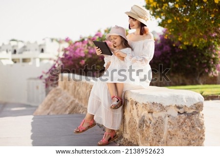 Happy modern mother and daughter in white dresses and straw hats near the hotel in a tropical resort using tablet PC while sitting on a background of a tree with pink flowers. Foto stock © 