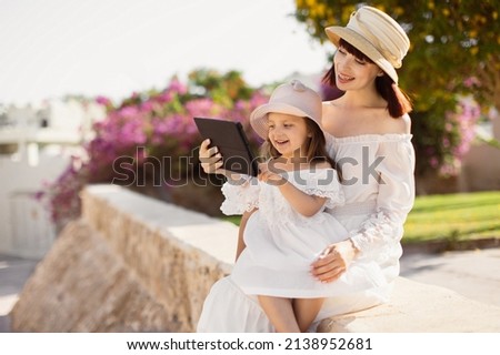 Happy modern mother and daughter in white dresses and straw hats near the hotel in a tropical resort using tablet PC while sitting on a background of a tree with pink flowers. Foto stock © 