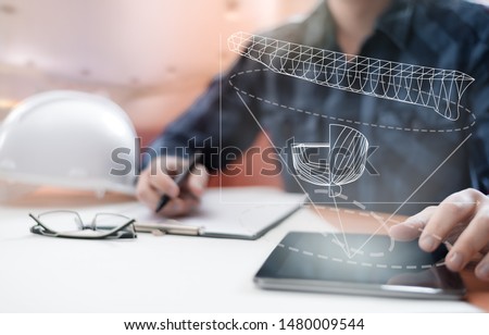 Young blurred sub-designer sitting in the office designing ship for future construction, with digital drawing on tablet PC and paper sketch. Ship design, construction and vessel architecture concept.