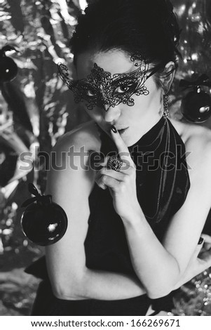 Mysterious young woman in black venetian mask and silk elegant dress showing a sign of silence. Unusual christmas decorations