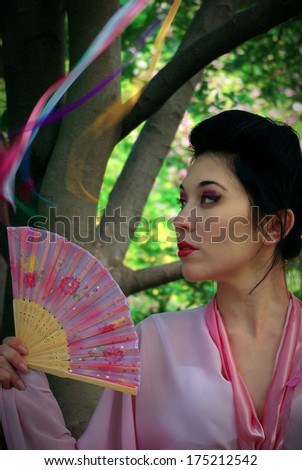 Portrait of Japanese young beautiful woman wearing  pink kimono with fan standing near the tree looking into the distance. Traditional makeup. Colorful silk ribbons fluttering  in the wind