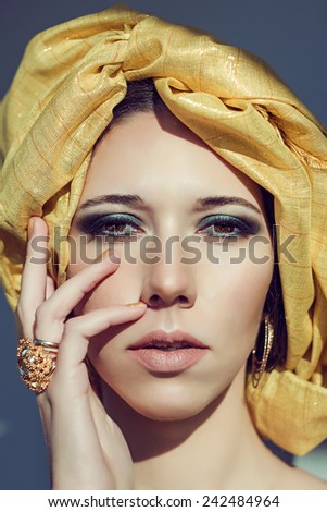 Close up fashion portrait of cryptic beautiful young arab woman in yellow turban. Gold jewelry necklace and  openwork ring with precious stones. Diamonds. Makeup smoky turquoise eyes. Model shooting.