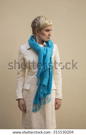 Stylish beautiful young woman blonde wearing blue scarf and white pattern coat isolated on beige background. Fashion haircut. Professional nude fresh makeup. Model shooting. Elegant style.