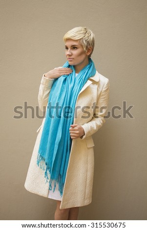 Stylish beautiful young woman blonde wearing blue scarf and white pattern coat isolated on beige background. fashion haircut. Professional nude fresh makeup. Model shooting. Elegant style.