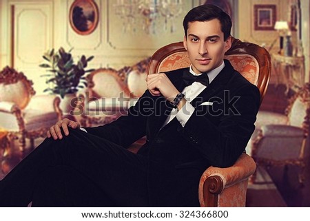 Confident successful young handsome man businessman in elegant suit with bow tie sitting on vintage armchair in luxurious living room. Manhood. Male beauty. Fashion model studio shot. Italian style.