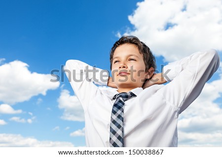 Boy in white shirt and a tie, his hands behind his back, he is confidently and happy looking into the distance. He is outside, with background a beautiful blue sky and white clouds