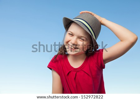 A portrait of a cute little girl, she is standing outside, wearing a hat and a red shirt against a blue sky,she is laughing