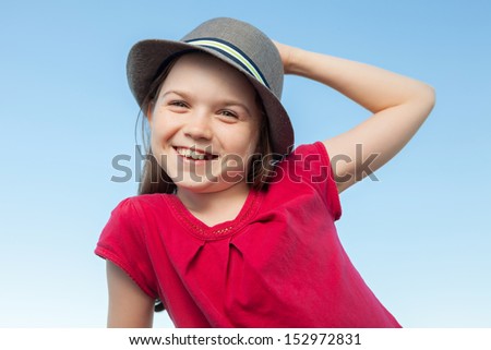 A portrait of a cute little girl, she is standing outside, wearing a hat and a red shirt against a blue sky,she is smiling into the camera