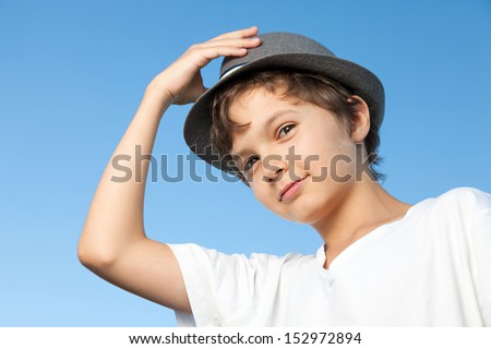 A portrait of a teenage boy, he is standing outside, wearing a white shirt against a blue sky. He is also wearing a hat, one hand is up at the hat, he is looking straight into the camera.