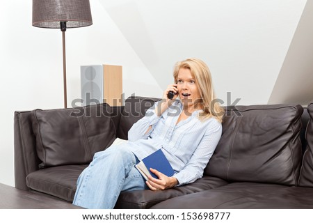 A beautiful blond woman is sitting on a couch, talking into the phone, having a conversation. She looks very surprised