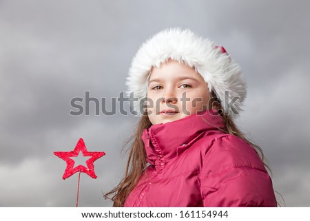 Cute young girl standing outside, wearing a red Santa hat,looking into the camera, holding a red Christmas star in her hands