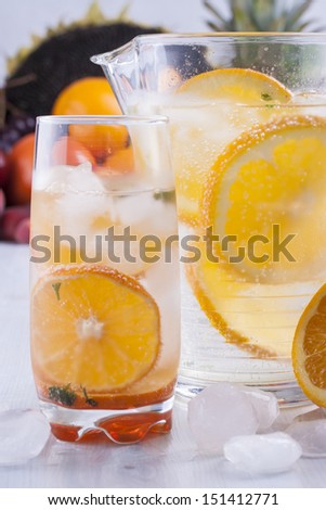 Fresh fruit and water drink with a orange slices, mint herb and ice cubes with some fruits in the background.