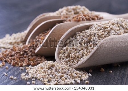 Close up photo of a raw eco food in the wooden spatula - light brown buckwheat groats, dark brown kasha and brown pearl barley placed on a dark wooden background.