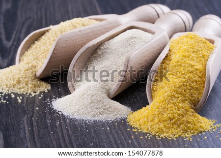 Close up photo of a raw eco food in the wooden spatulas - light yellow couscous, light brown farina and dark yellow cornmeal placed on a dark wooden background.