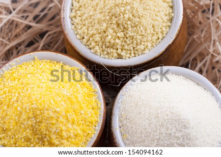 Close up photo of a raw eco food in the clay cup - light yellow couscous, light brown farina and dark yellow cornmeal placed on a dark wooden background.