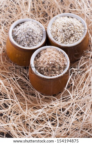 Close up photo of a cereal grain product in a clay cup - dark brown wheat bran, light brown secale bran, and mid-brown avena bran placed on a wooden shavings.