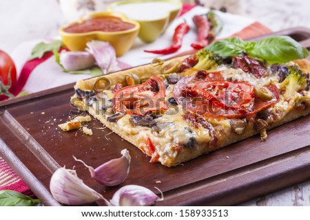 Close up of the just baked homemade pizza with tomato, salami slices and olives placed on brown cutting board.