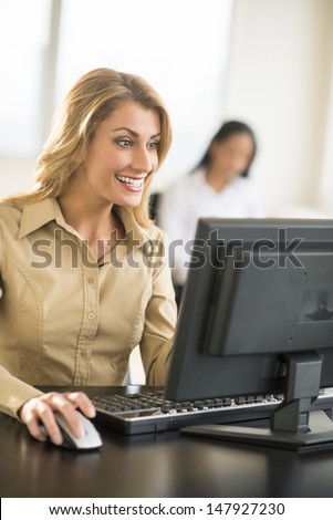 Happy young businesswoman using computer at desk in office with female colleague sitting in background