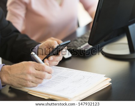 Midsection of businessman using smart phone while writing on document with colleague at desk in office