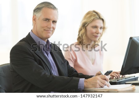 Portrait of confident mature businessman holding pen and mobile phone with female colleague using computer in office