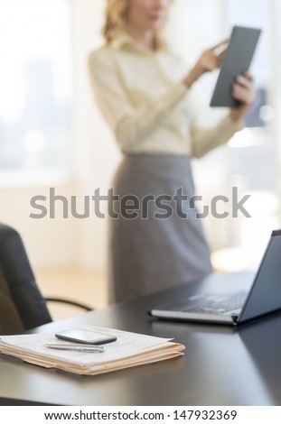 Documents and laptop on desk with businesswoman using digital tablet in background at office