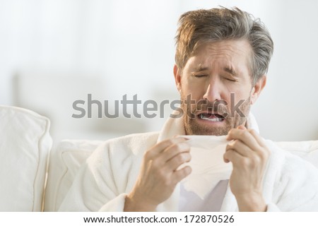 Mature man sneezing into tissue on sofa at home