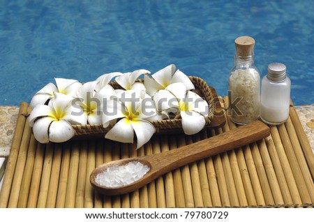 Bowl of frangipani and herbal salt in spoon and massage oil on mat in the swimming pool