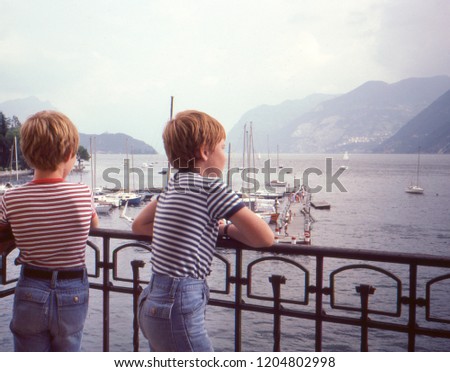 Vintage 1982 analog camera image of two young boys / brothers seen from behind both wearing jeans shorts and striped t-shirts overlooking lake Geneva or Lac Léman from a balcony in Switzerland. Imagine de stoc © 