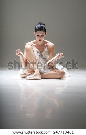 Young beautiful ballerina sitting under spotlight in costumed tutu adjusting pointed ballet shoe strapping