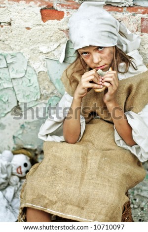 Sad child wearing vintage, dirty clothing, with a piece of bread in her hand and smiling doll. More available.