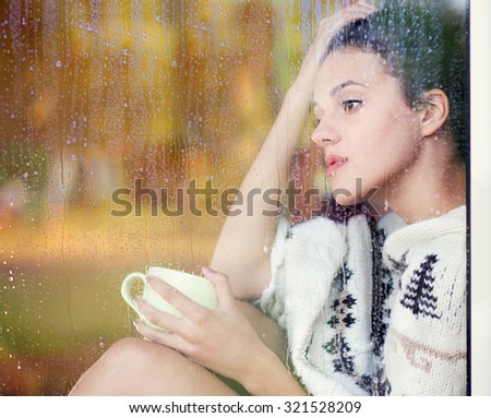 Beautiful thoughtful brunette woman with cup of coffee wearing knitted nordic print poncho sitting home by the window covered with rain drops. Blurred garden fall background reflection.
