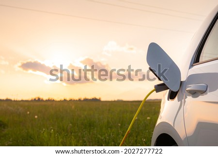 Electric car with opened charging socket cap and charger plugged in, at a public electric charging station near the highway at sunset