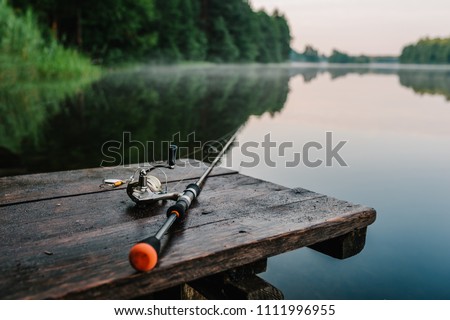 Fishing rod, spinning reel on the background pier river bank. Sunrise. Fog against the backdrop of lake. Misty morning. wild nature. The concept of rural getaway. Article about fishing day.