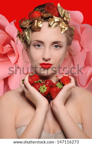portrait of a beautiful blonde girl with strawberries in hands and on her head a crown of gold leaf on a red background
