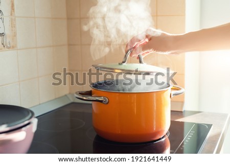 Female hand open lid of enamel steel cooking pan on electric hob with boiling water or soup and scenic vapor steam backlit by warm sunlight at kitchen. Kitchenware utensil and tool at home background
