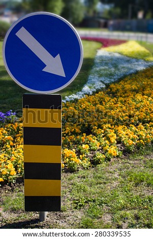 Photo of traffic sign for mandatory direction that is placed on the lawn in front of flower garden full of flowers in orange, white, yellow and red color