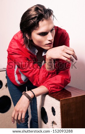 Young handsome male in red jacket and blue jeans with beautiful face, hairstyle, jewelry and his hands on speakers looking down