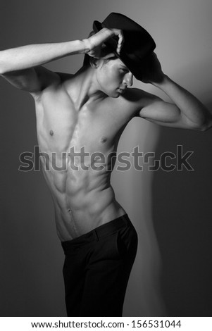Sensual young guy in hat with beautiful face and muscular torso, with hands on hat, dressed in black trousers and looking away Black and White Photo