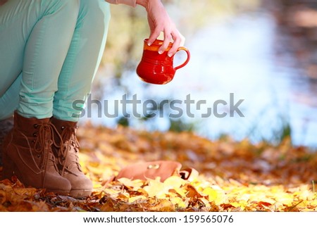 Woman relaxing in the autumn park enjoying hot drink coffee or tea, female hand holding red mug with warm beverage. Yellow leaves background