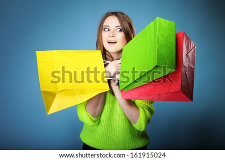 Surprised emotional woman in bright vivid colour sweater with paper shopping bags on blue background. Sales and discounts concept.